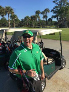 Smiling man in a green polo shirt with a golf cart and clubs on a sunny golf course background with palm trees behind him.